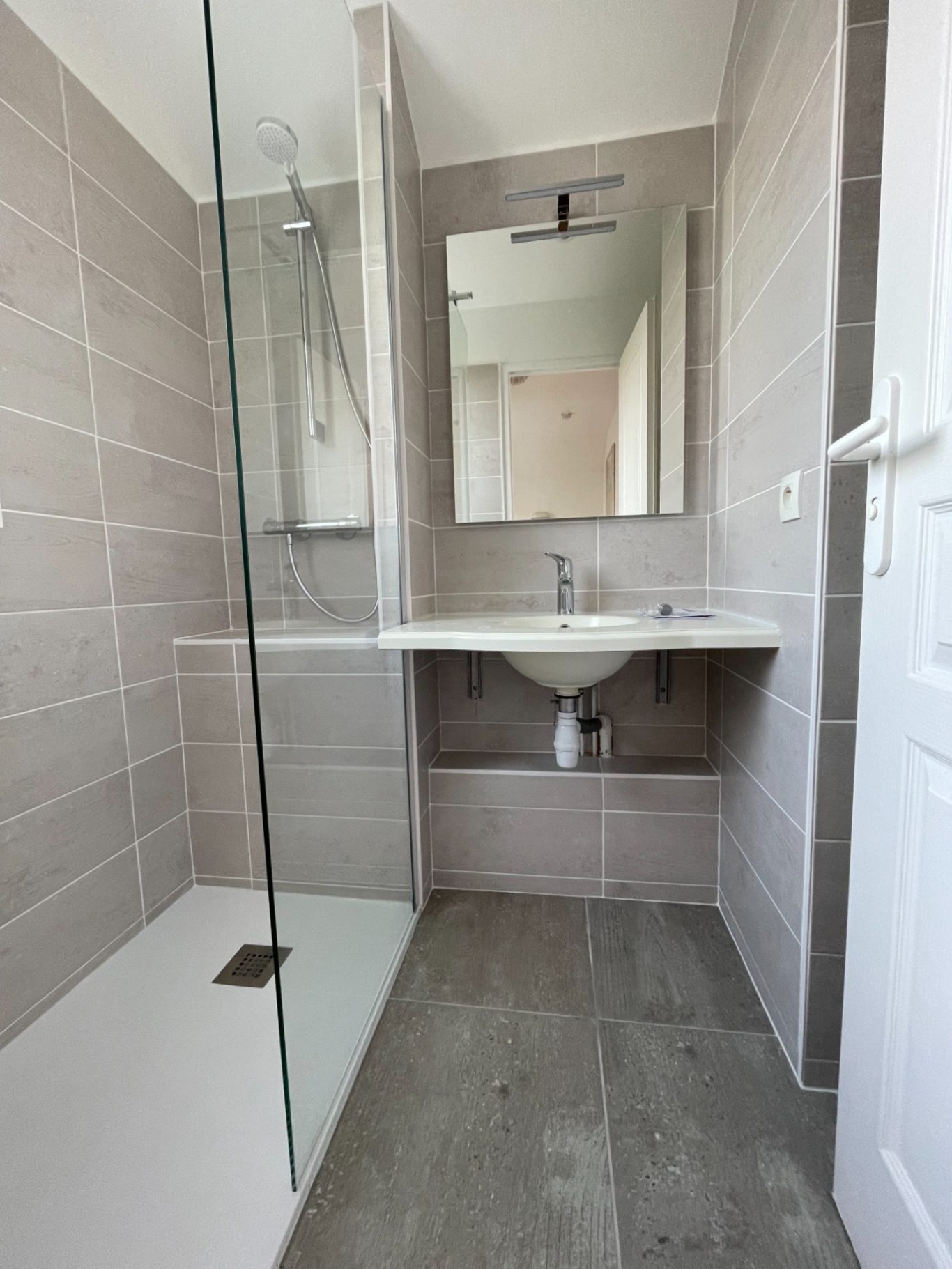 Salle de bain moderne avec douche, lavabo et miroir sur un mur carrelé clair.