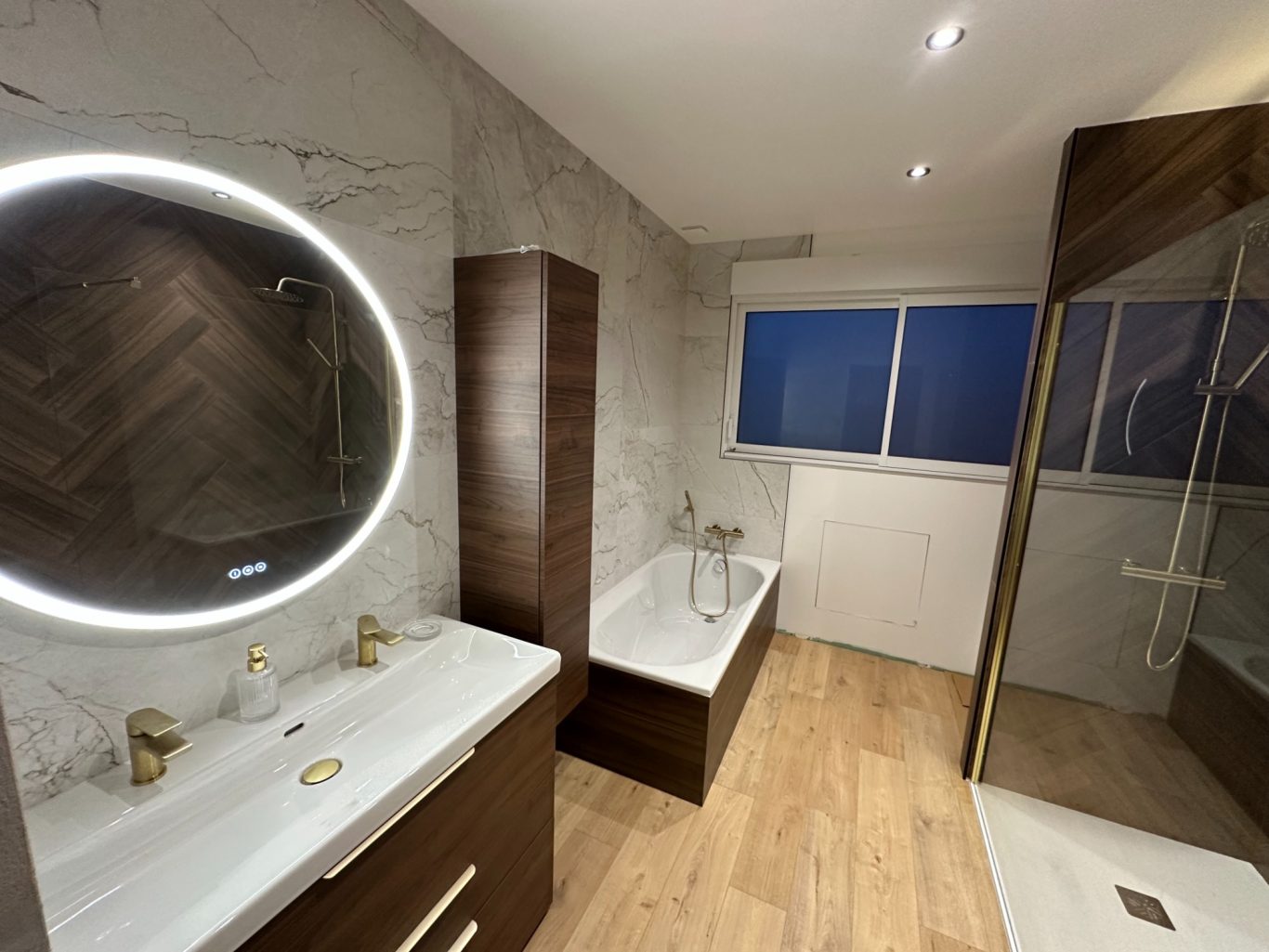 Salle de bains moderne avec miroir rond, baignoire, douche et meubles en bois.