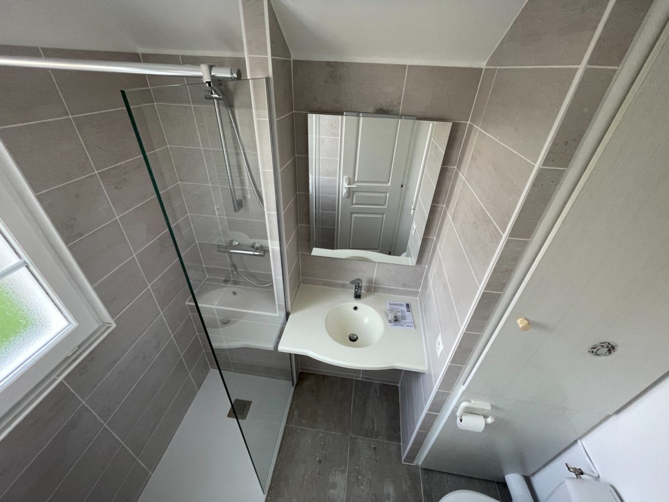 Salle de bain moderne avec douche, lavabo et miroir en coin. Mur en carrelage gris.