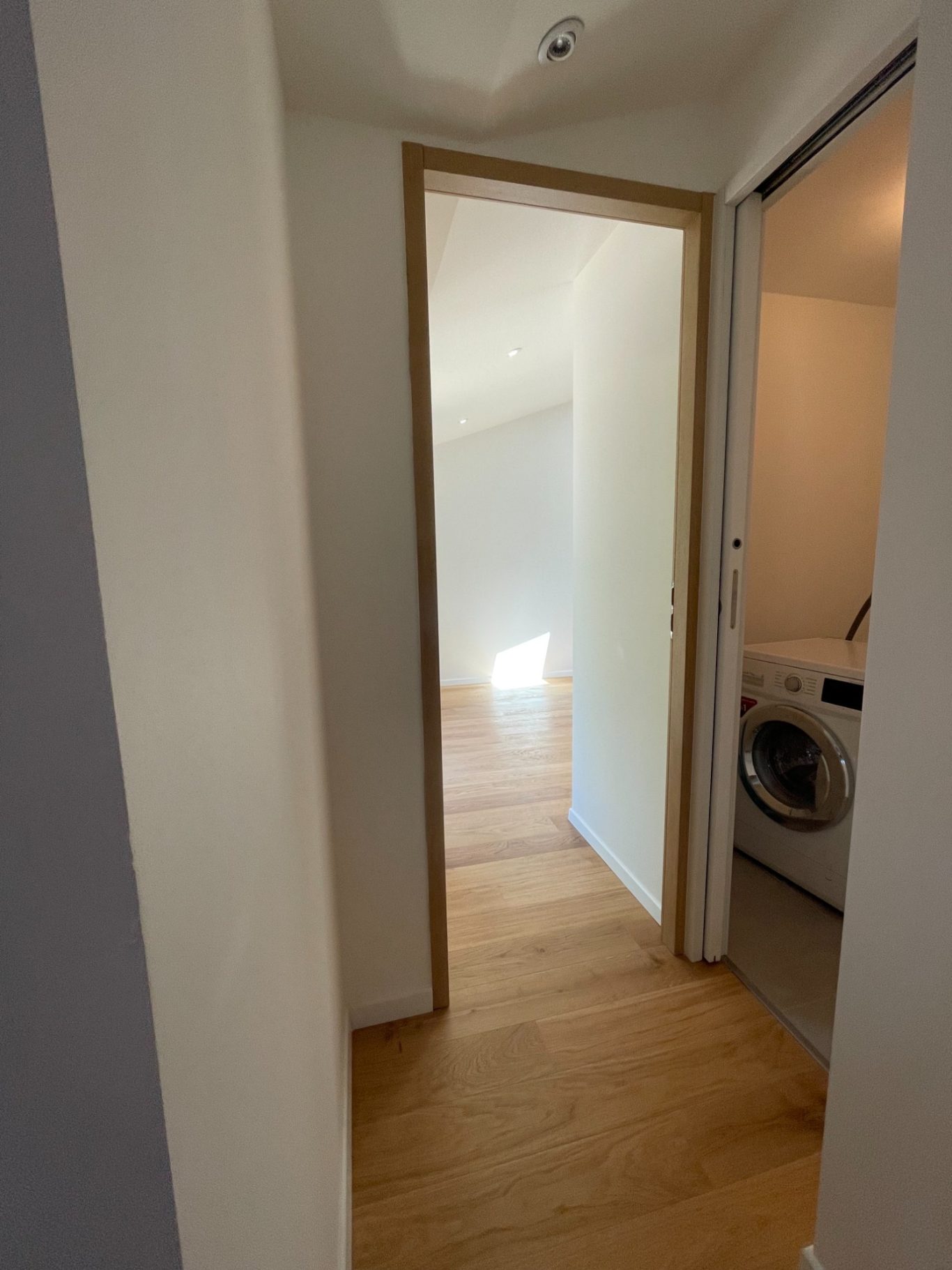 Un couloir lumineux menant à une pièce avec des murs clairs et un lave-linge visible sur la droite.